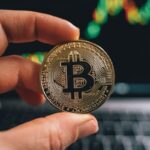 Close-up of a hand holding a Bitcoin coin in front of a trading screen, symbolizing cryptocurrency trading.