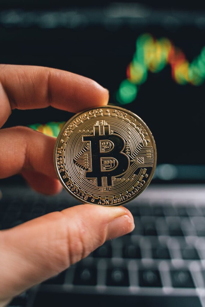 Close-up of a hand holding a Bitcoin coin in front of a trading screen, symbolizing cryptocurrency trading.